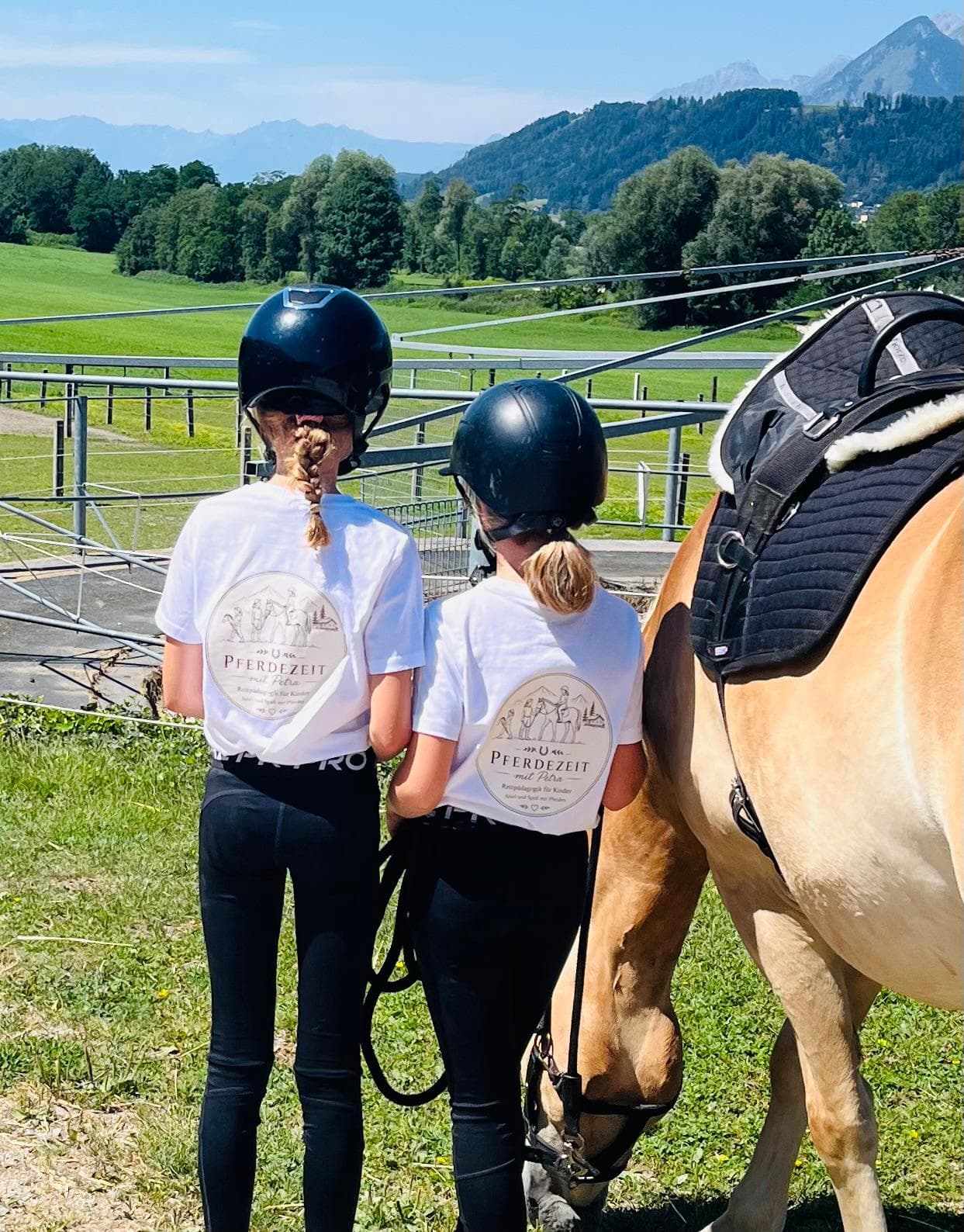 Zwei Kinder mit Reithelmen stehen mit einem Haflinger am Zaun und blicken ins Inntal